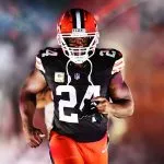 Nick Chubb, ein schwarzes Cleveland Browns-Trikot mit der Nummer 24 und orangefarbene Hosen, rennt auf das Spielfeld. Er hält seinen Mundschutz, während grauer Rauch und bunte Lichter im Hintergrund für einen dramatischen Auftritt sorgen. Diese Beschreibung wurde mit der FootballR KI automatisch generiert.