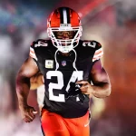 Nick Chubb, ein schwarzes Cleveland Browns-Trikot mit der Nummer 24 und orangefarbene Hosen, rennt auf das Spielfeld. Er hält seinen Mundschutz, während grauer Rauch und bunte Lichter im Hintergrund für einen dramatischen Auftritt sorgen. Diese Beschreibung wurde mit der FootballR KI automatisch generiert.