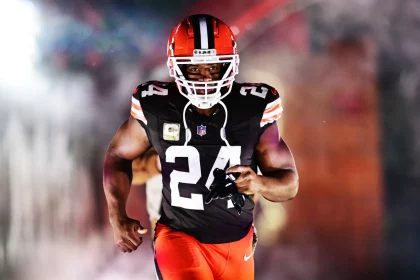 Nick Chubb, ein schwarzes Cleveland Browns-Trikot mit der Nummer 24 und orangefarbene Hosen, rennt auf das Spielfeld. Er hält seinen Mundschutz, während grauer Rauch und bunte Lichter im Hintergrund für einen dramatischen Auftritt sorgen. Diese Beschreibung wurde mit der FootballR KI automatisch generiert.