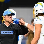 Ein Chargers-Trainer in einem schwarzen Kapuzenpulli und einer blauen Mütze stößt Justin Herbert, der eine weiße Chargers-Uniform und einen Helm mit der Nummer 10 trägt, mit der Faust an. Der unscharfe Hintergrund lenkt den Fokus auf ihre Jubelgeste. Diese Beschreibung wurde mit der FootballR KI automatisch generiert.