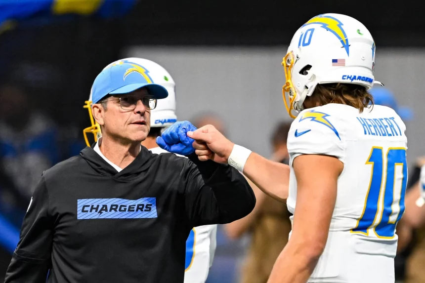 Ein Chargers-Trainer in einem schwarzen Kapuzenpulli und einer blauen Mütze stößt Justin Herbert, der eine weiße Chargers-Uniform und einen Helm mit der Nummer 10 trägt, mit der Faust an. Der unscharfe Hintergrund lenkt den Fokus auf ihre Jubelgeste. Diese Beschreibung wurde mit der FootballR KI automatisch generiert.