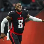 Kyle Pitts trägt eine schwarze Uniform der Atlanta Falcons mit der Nummer 8, weißen Ärmeln und roten Akzenten und läuft mit ausgestrecktem Arm über das Spielfeld. Der Hintergrund ist verschwommen, mit undeutlichen Figuren und Stadionlichtern. Diese Beschreibung wurde mit der FootballR KI automatisch generiert.