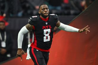 Kyle Pitts trägt eine schwarze Uniform der Atlanta Falcons mit der Nummer 8, weißen Ärmeln und roten Akzenten und läuft mit ausgestrecktem Arm über das Spielfeld. Der Hintergrund ist verschwommen, mit undeutlichen Figuren und Stadionlichtern. Diese Beschreibung wurde mit der FootballR KI automatisch generiert.