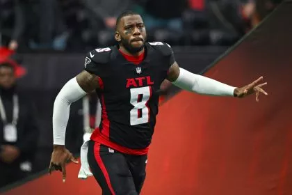 Kyle Pitts trägt eine schwarze Uniform der Atlanta Falcons mit der Nummer 8, weißen Ärmeln und roten Akzenten und läuft mit ausgestrecktem Arm über das Spielfeld. Der Hintergrund ist verschwommen, mit undeutlichen Figuren und Stadionlichtern. Diese Beschreibung wurde mit der FootballR KI automatisch generiert.