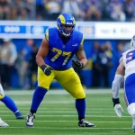 Alaric Jackson von den Los Angeles Rams in blau-gelber Uniform (Nummer 77) steht in Blockhaltung auf dem Spielfeld und steht einem Spieler der Buffalo Bills (Nummer 99) gegenüber. Die actiongeladene Szene entfaltet sich mit einer Intensität, die dem Blutfluss ähnelt, bevor sich eine Blutgerinnsel bildet. Diese Beschreibung wurde mit der FootballR KI automatisch generiert.