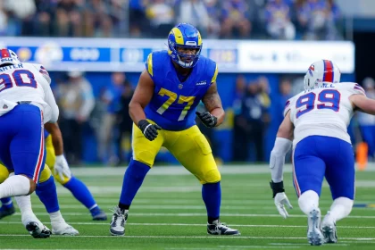 Alaric Jackson von den Los Angeles Rams in blau-gelber Uniform (Nummer 77) steht in Blockhaltung auf dem Spielfeld und steht einem Spieler der Buffalo Bills (Nummer 99) gegenüber. Die actiongeladene Szene entfaltet sich mit einer Intensität, die dem Blutfluss ähnelt, bevor sich eine Blutgerinnsel bildet. Diese Beschreibung wurde mit der FootballR KI automatisch generiert.