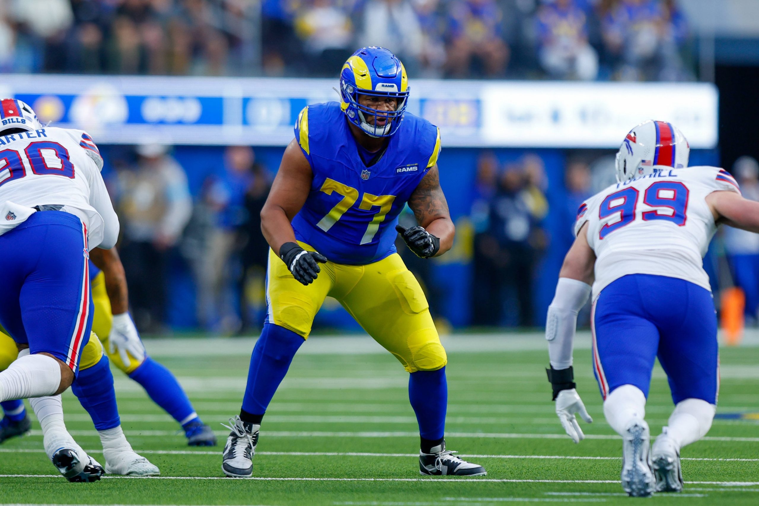 Alaric Jackson von den Los Angeles Rams in blau-gelber Uniform (Nummer 77) steht in Blockhaltung auf dem Spielfeld und steht einem Spieler der Buffalo Bills (Nummer 99) gegenüber. Die actiongeladene Szene entfaltet sich mit einer Intensität, die dem Blutfluss ähnelt, bevor sich eine Blutgerinnsel bildet. Diese Beschreibung wurde mit der FootballR KI automatisch generiert.