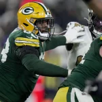 Pass Protection - Ein Footballspieler der Green Bay Packers in grünem Trikot und gelbem Helm blockt während eines Spiels einen gegnerischen Spieler in Weiß. Der Packers-Spieler streckt seinen Arm aus und greift körperlich an, während andere Spieler im Hintergrund teilweise sichtbar sind. Diese Beschreibung wurde mit der FootballR KI automatisch generiert.