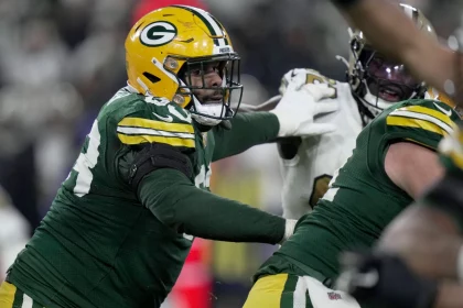 Pass Protection - Ein Footballspieler der Green Bay Packers in grünem Trikot und gelbem Helm blockt während eines Spiels einen gegnerischen Spieler in Weiß. Der Packers-Spieler streckt seinen Arm aus und greift körperlich an, während andere Spieler im Hintergrund teilweise sichtbar sind. Diese Beschreibung wurde mit der FootballR KI automatisch generiert.