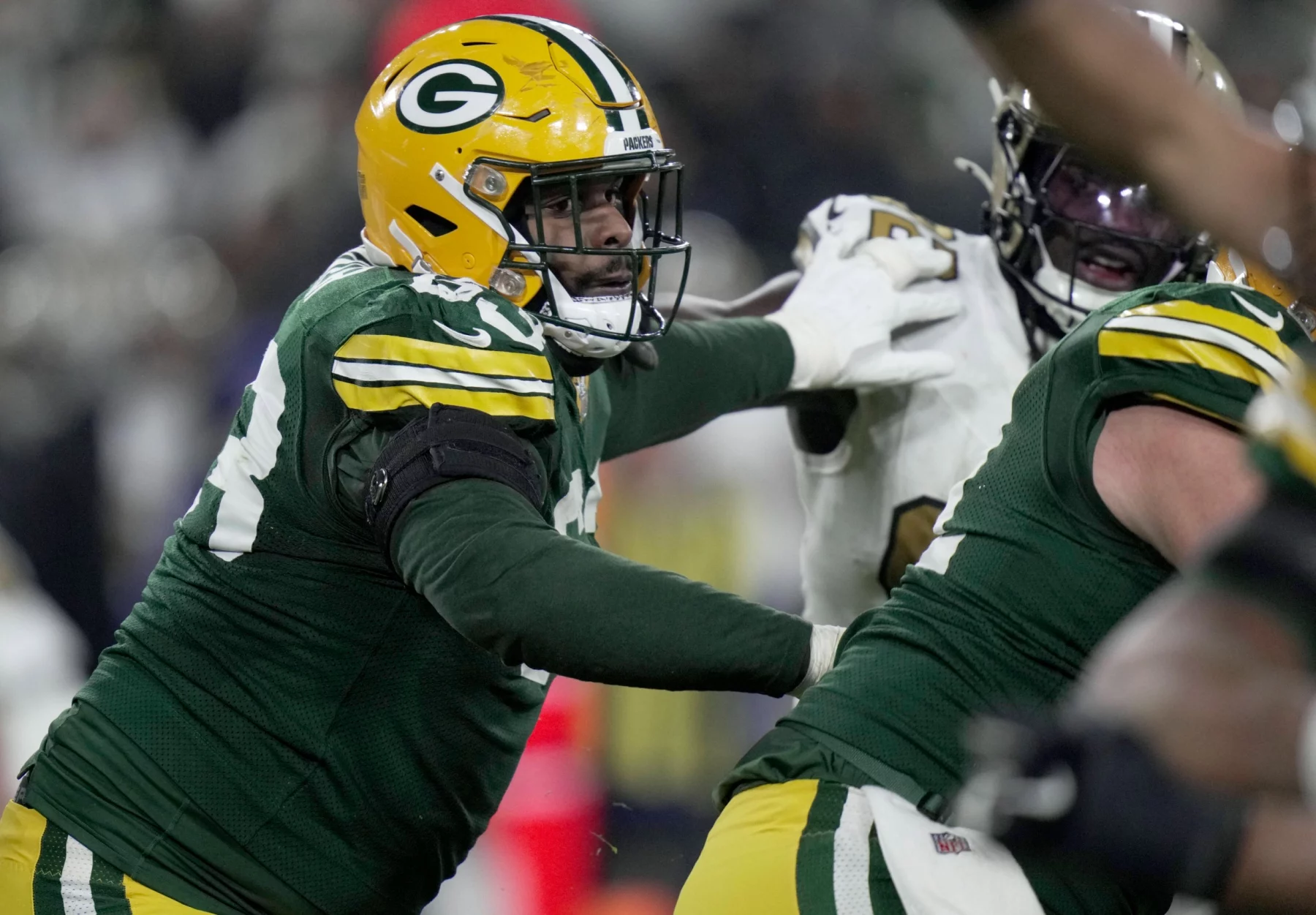 Pass Protection - Ein Footballspieler der Green Bay Packers in grünem Trikot und gelbem Helm blockt während eines Spiels einen gegnerischen Spieler in Weiß. Der Packers-Spieler streckt seinen Arm aus und greift körperlich an, während andere Spieler im Hintergrund teilweise sichtbar sind. Diese Beschreibung wurde mit der FootballR KI automatisch generiert.