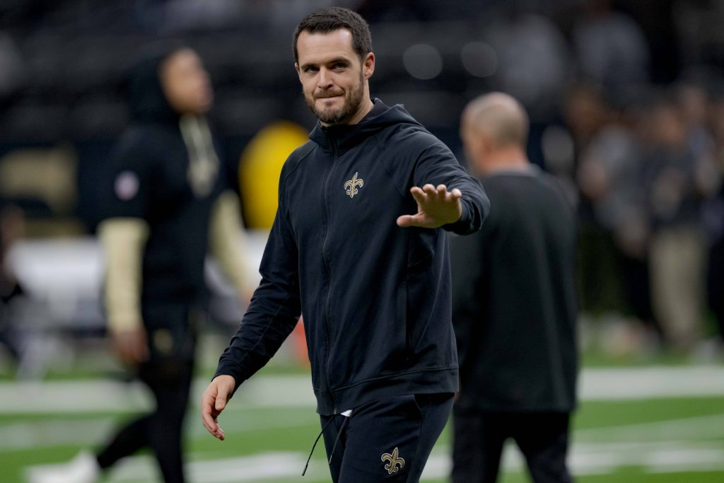 Derek Carr im schwarzen Trainingsanzug der New Orleans Saints, der Derek Carr ähnelt, lächelt und winkt auf einem Footballfeld. Der unscharfe Hintergrund hebt das Stadion und die Menge hervor und lenkt den Fokus auf die fröhliche Figur im Vordergrund. Diese Beschreibung wurde mit der FootballR KI automatisch generiert.