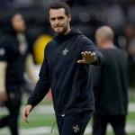 Derek Carr im schwarzen Trainingsanzug der New Orleans Saints, der Derek Carr ähnelt, lächelt und winkt auf einem Footballfeld. Der unscharfe Hintergrund hebt das Stadion und die Menge hervor und lenkt den Fokus auf die fröhliche Figur im Vordergrund. Diese Beschreibung wurde mit der FootballR KI automatisch generiert.