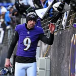 Der NFL-Spieler Justin Tucker von den Baltimore Ravens, der ein lila Trikot mit der Nummer 9 und eine Mütze trägt, gibt den Fans über das Stadiongeländer ein High Five. Er hält seinen Helm in einer Hand und blickt nach oben, während die Menge Fahnen schwenkt und sich begeistert die Hände reicht. Diese Beschreibung wurde mit der FootballR KI automatisch generiert.