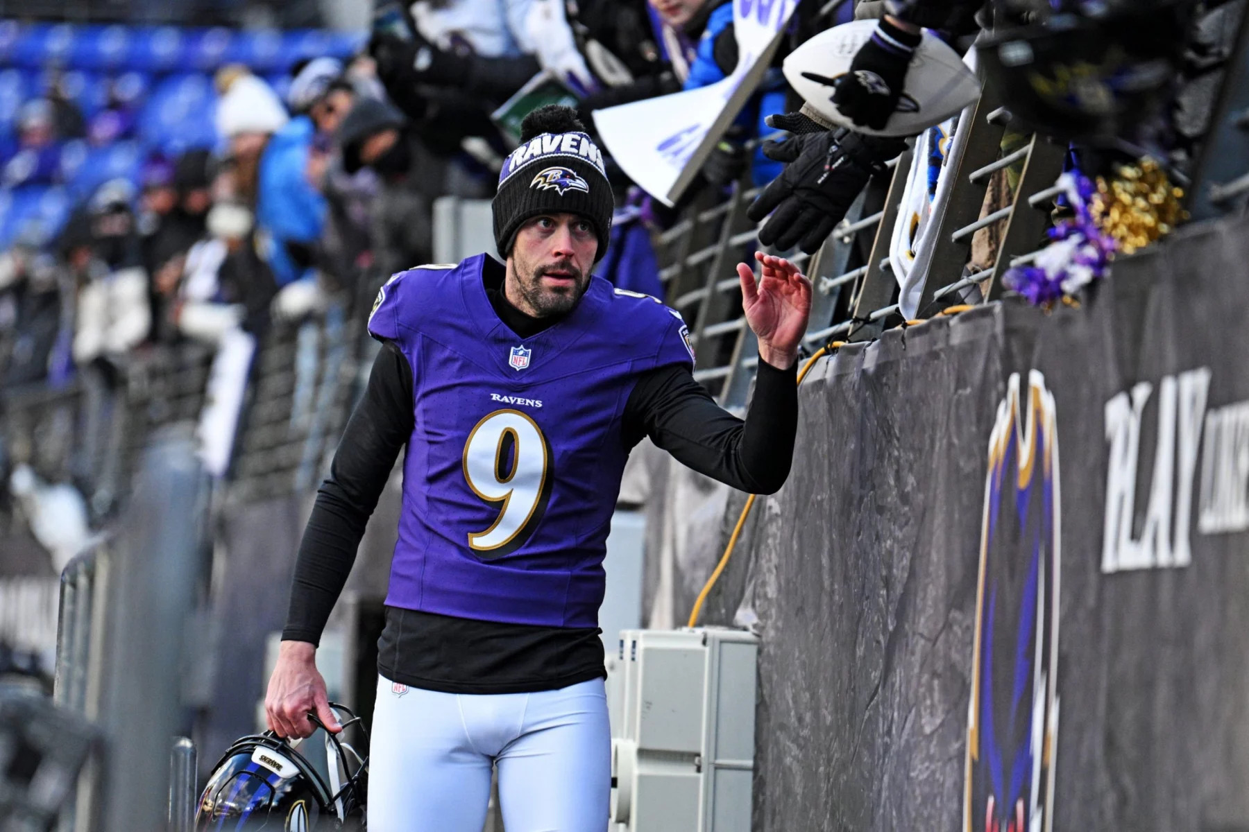Der NFL-Spieler Justin Tucker von den Baltimore Ravens, der ein lila Trikot mit der Nummer 9 und eine Mütze trägt, gibt den Fans über das Stadiongeländer ein High Five. Er hält seinen Helm in einer Hand und blickt nach oben, während die Menge Fahnen schwenkt und sich begeistert die Hände reicht. Diese Beschreibung wurde mit der FootballR KI automatisch generiert.