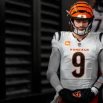 Ein konzentrierter Joe Burrow, in einer weißen Uniform der Cincinnati Bengals mit orange-schwarzem Helm (Nummer 9), steht mit gefalteten Händen in einem dunklen Tunnel. Andere Spieler sind leicht zu erkennen, während er sich auf das Spiel vorbereitet. Diese Beschreibung wurde mit der FootballR KI automatisch generiert.