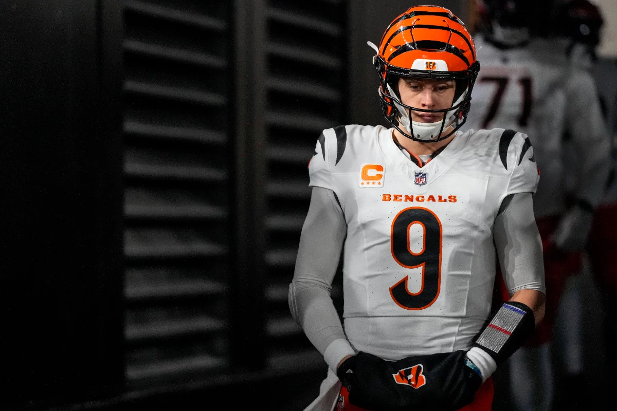 Ein konzentrierter Joe Burrow, in einer weißen Uniform der Cincinnati Bengals mit orange-schwarzem Helm (Nummer 9), steht mit gefalteten Händen in einem dunklen Tunnel. Andere Spieler sind leicht zu erkennen, während er sich auf das Spiel vorbereitet. Diese Beschreibung wurde mit der FootballR KI automatisch generiert.