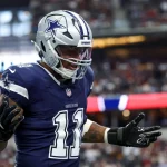 Dallas Cowboys-Star Micah Parsons, Trikotnummer 11, silberner Helm und schwarze Handschuhe, steht mit erhobenen Händen und ausdrucksstarkem Gesicht auf dem Spielfeld. Im Hintergrund leuchten die Zuschauer und die Stadionlichter unscharf. Diese Beschreibung wurde mit der FootballR KI automatisch generiert.