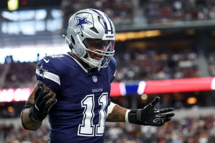 Dallas Cowboys-Star Micah Parsons, Trikotnummer 11, silberner Helm und schwarze Handschuhe, steht mit erhobenen Händen und ausdrucksstarkem Gesicht auf dem Spielfeld. Im Hintergrund leuchten die Zuschauer und die Stadionlichter unscharf. Diese Beschreibung wurde mit der FootballR KI automatisch generiert.