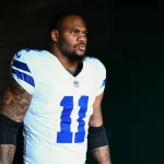NFC East - Ein Footballspieler, Micah Parsons, trägt ein weißes Dallas Cowboys-Trikot mit der Nummer 11 und steht im Halbdunkel. Er hat einen ernsten Gesichtsausdruck, sichtbare Armtattoos und schwarze Kompressionsstrümpfe. Der dunkle Hintergrund unterstreicht seine Präsenz. Diese Beschreibung wurde mit der FootballR KI automatisch generiert.