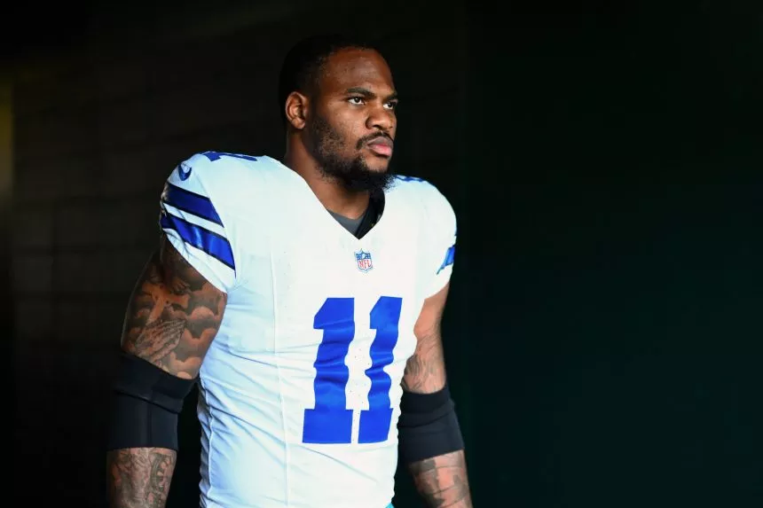 Ein Footballspieler, Micah Parsons, trägt ein weißes Dallas Cowboys-Trikot mit der Nummer 11 und steht im Halbdunkel. Er hat einen ernsten Gesichtsausdruck, sichtbare Armtattoos und schwarze Kompressionsstrümpfe. Der dunkle Hintergrund unterstreicht seine Präsenz. Diese Beschreibung wurde mit der FootballR KI automatisch generiert.