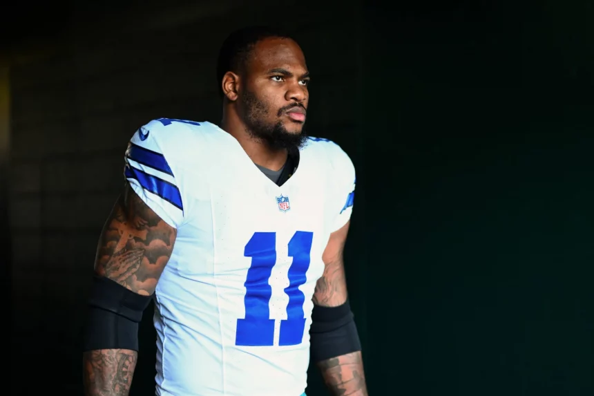 NFC East - Ein Footballspieler, Micah Parsons, trägt ein weißes Dallas Cowboys-Trikot mit der Nummer 11 und steht im Halbdunkel. Er hat einen ernsten Gesichtsausdruck, sichtbare Armtattoos und schwarze Kompressionsstrümpfe. Der dunkle Hintergrund unterstreicht seine Präsenz. Diese Beschreibung wurde mit der FootballR KI automatisch generiert.
