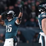 Saquon Barkley, ein Footballspieler der Philadelphia Eagles, mit der Trikotnummer 26 jubelt mit einem Daumen-hoch-Geste auf dem Spielfeld und sieht aus, als wäre er bereit für das Madden NFL 26 Cover. Er steht in voller Uniform neben einem Teamkollegen, im Hintergrund sind Fans und Stadionlichter verschwommen. Diese Beschreibung wurde mit der FootballR KI automatisch generiert.