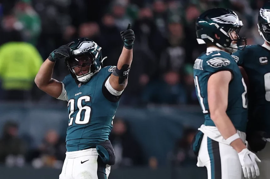 Saquon Barkley, ein Footballspieler der Philadelphia Eagles, mit der Trikotnummer 26 jubelt mit einem Daumen-hoch-Geste auf dem Spielfeld und sieht aus, als wäre er bereit für das Madden NFL 26 Cover. Er steht in voller Uniform neben einem Teamkollegen, im Hintergrund sind Fans und Stadionlichter verschwommen. Diese Beschreibung wurde mit der FootballR KI automatisch generiert.