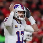 Ein Football-Spieler, Josh Allen, in einer weißen Buffalo Bills Uniform und Helm, Nummer 17, steht auf dem Spielfeld mit beiden Händen an seinem Helm und wirkt konzentriert oder animiert; eine verschwommene Menge in Rot füllt den Hintergrund. Diese Beschreibung wurde mit der FootballR KI automatisch generiert.