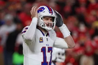 Ein Football-Spieler, Josh Allen, in einer weißen Buffalo Bills Uniform und Helm, Nummer 17, steht auf dem Spielfeld mit beiden Händen an seinem Helm und wirkt konzentriert oder animiert; eine verschwommene Menge in Rot füllt den Hintergrund. Diese Beschreibung wurde mit der FootballR KI automatisch generiert.