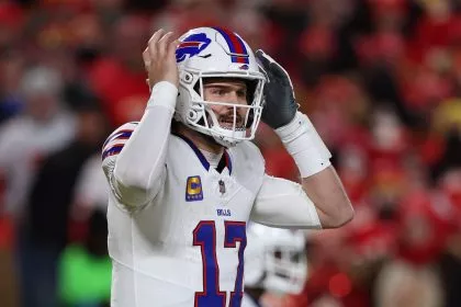 Ein Football-Spieler, Josh Allen, in einer weißen Buffalo Bills Uniform und Helm, Nummer 17, steht auf dem Spielfeld mit beiden Händen an seinem Helm und wirkt konzentriert oder animiert; eine verschwommene Menge in Rot füllt den Hintergrund. Diese Beschreibung wurde mit der FootballR KI automatisch generiert.