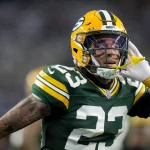 Jaire Alexander, Star der Green Bay Packers, trägt das Trikot mit der Nummer 23 und einen gelben Helm und führt seine linke Hand mit offenem Mundschutz ans Ohr. Der Hintergrund ist verschwommen, was Alexanders ausdrucksstarke Geste und sein konzentriertes Gesicht hervorhebt. Diese Beschreibung wurde mit der FootballR KI automatisch generiert.