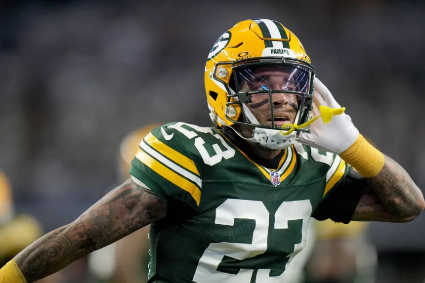Jaire Alexander, Star der Green Bay Packers, trägt das Trikot mit der Nummer 23 und einen gelben Helm und führt seine linke Hand mit offenem Mundschutz ans Ohr. Der Hintergrund ist verschwommen, was Alexanders ausdrucksstarke Geste und sein konzentriertes Gesicht hervorhebt. Diese Beschreibung wurde mit der FootballR KI automatisch generiert.
