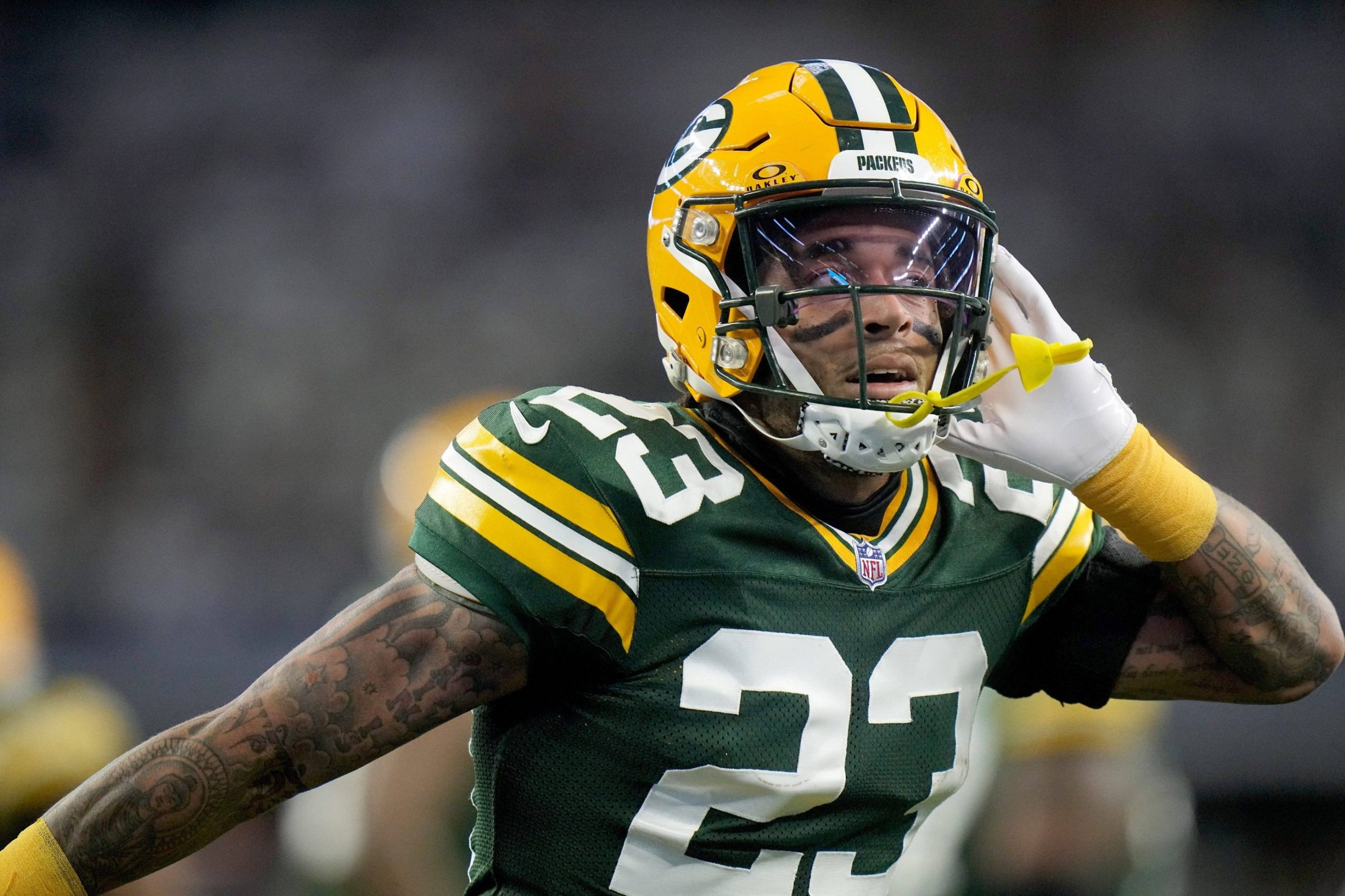 Jaire Alexander, Star der Green Bay Packers, trägt das Trikot mit der Nummer 23 und einen gelben Helm und führt seine linke Hand mit offenem Mundschutz ans Ohr. Der Hintergrund ist verschwommen, was Alexanders ausdrucksstarke Geste und sein konzentriertes Gesicht hervorhebt. Diese Beschreibung wurde mit der FootballR KI automatisch generiert.