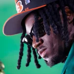 Shemar Stewart, schwarzen Dreadlocks und Sonnenbrille trägt eine schwarz-orange Bengals-Kappe und einen grünen Blazer. Ihr Gesicht ist vor einem hellgrünen Hintergrund nach unten geneigt, was ihr einen ernsten Ausdruck verleiht. Goldene Creolen und Gesichtsbehaarung sind sichtbar. Diese Beschreibung wurde mit der FootballR KI automatisch generiert.