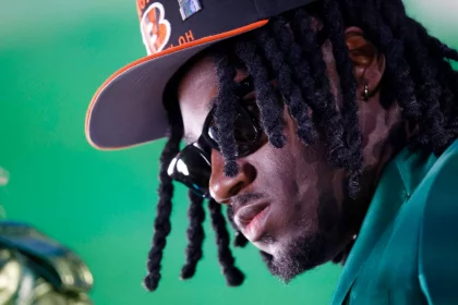 Shemar Stewart, schwarzen Dreadlocks und Sonnenbrille trägt eine schwarz-orange Bengals-Kappe und einen grünen Blazer. Ihr Gesicht ist vor einem hellgrünen Hintergrund nach unten geneigt, was ihr einen ernsten Ausdruck verleiht. Goldene Creolen und Gesichtsbehaarung sind sichtbar. Diese Beschreibung wurde mit der FootballR KI automatisch generiert.