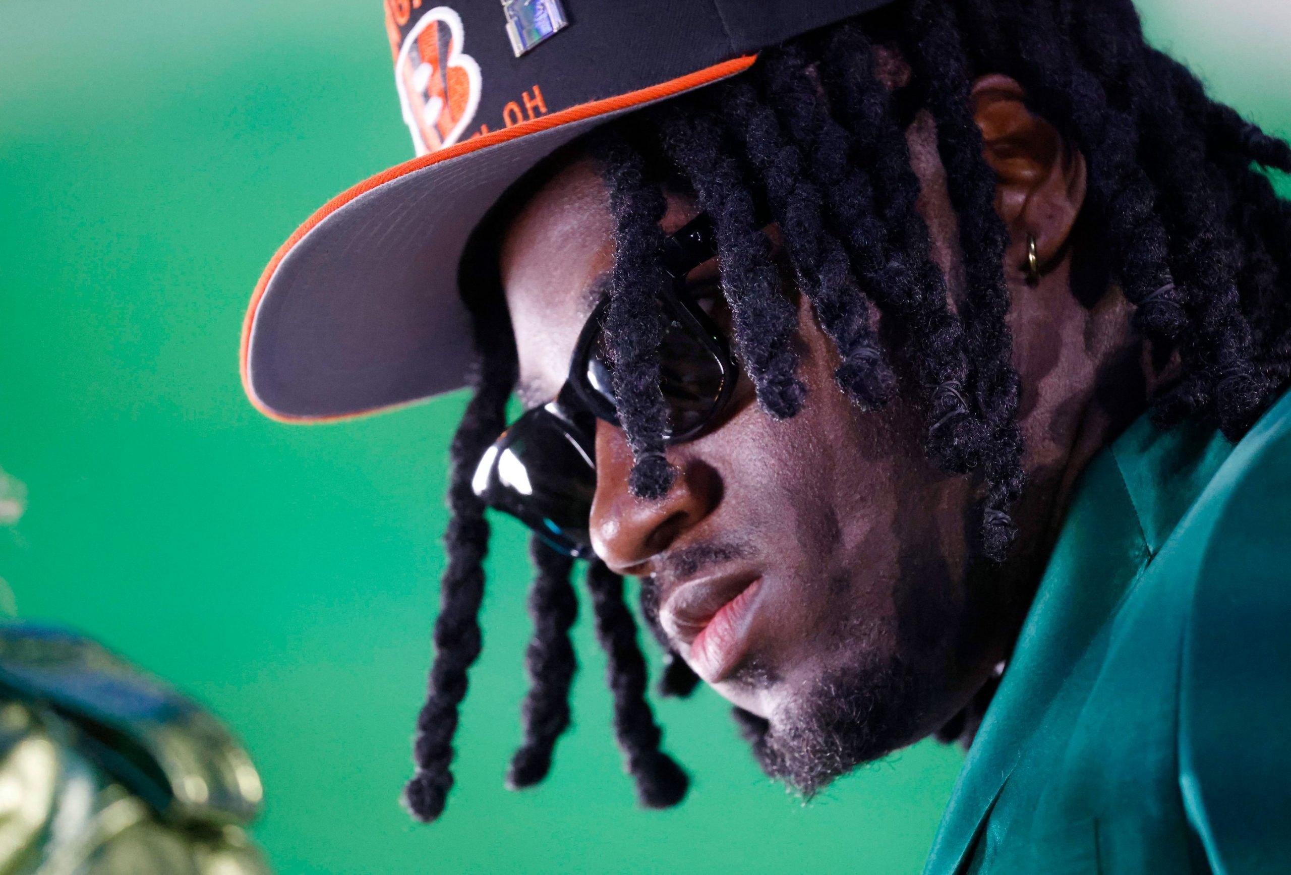 Shemar Stewart, schwarzen Dreadlocks und Sonnenbrille trägt eine schwarz-orange Bengals-Kappe und einen grünen Blazer. Ihr Gesicht ist vor einem hellgrünen Hintergrund nach unten geneigt, was ihr einen ernsten Ausdruck verleiht. Goldene Creolen und Gesichtsbehaarung sind sichtbar. Diese Beschreibung wurde mit der FootballR KI automatisch generiert.