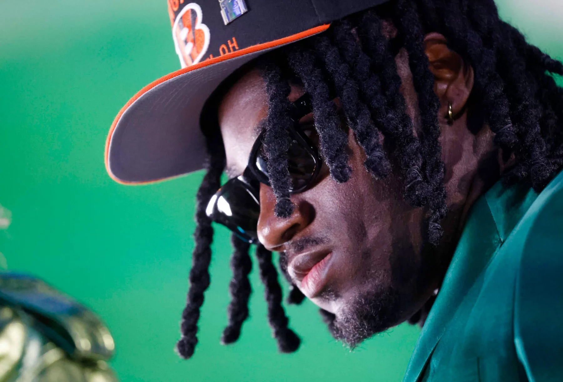 Shemar Stewart, schwarzen Dreadlocks und Sonnenbrille trägt eine schwarz-orange Bengals-Kappe und einen grünen Blazer. Ihr Gesicht ist vor einem hellgrünen Hintergrund nach unten geneigt, was ihr einen ernsten Ausdruck verleiht. Goldene Creolen und Gesichtsbehaarung sind sichtbar. Diese Beschreibung wurde mit der FootballR KI automatisch generiert.