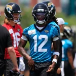 Travis Hunter trägt ein blaues Jacksonville Jaguars-Trikot mit der Nummer 12, einen schwarzen Helm und schwarze Handschuhe und steht beim Training im Freien. Travis Hunter ist unter den Spielern und Trainern in roten und weißen Hemden im unscharfen Hintergrund zu sehen. Diese Beschreibung wurde mit der FootballR KI automatisch generiert.