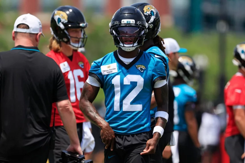 Travis Hunter trägt ein blaues Jacksonville Jaguars-Trikot mit der Nummer 12, einen schwarzen Helm und schwarze Handschuhe und steht beim Training im Freien. Travis Hunter ist unter den Spielern und Trainern in roten und weißen Hemden im unscharfen Hintergrund zu sehen. Diese Beschreibung wurde mit der FootballR KI automatisch generiert.