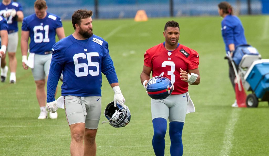 Zwei American-Football-Spieler laufen während des Giants Training auf einem Rasenplatz. Der linke Spieler trägt ein blaues Trikot mit der Nummer 65 und hält einen Helm; der rechte trägt ein rotes Trikot mit der Nummer 3 und trägt einen blauen Helm. Diese Beschreibung wurde mit der FootballR KI automatisch generiert.