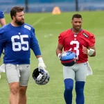 Zwei American-Football-Spieler laufen während des Giants Training auf einem Rasenplatz. Der linke Spieler trägt ein blaues Trikot mit der Nummer 65 und hält einen Helm; der rechte trägt ein rotes Trikot mit der Nummer 3 und trägt einen blauen Helm. Diese Beschreibung wurde mit der FootballR KI automatisch generiert.