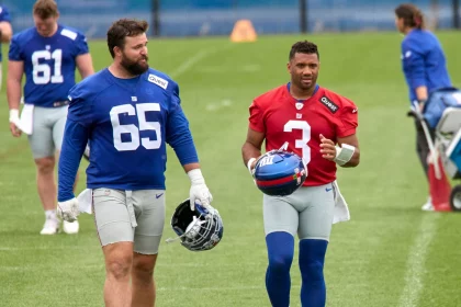 Zwei American-Football-Spieler laufen während des Giants Training auf einem Rasenplatz. Der linke Spieler trägt ein blaues Trikot mit der Nummer 65 und hält einen Helm; der rechte trägt ein rotes Trikot mit der Nummer 3 und trägt einen blauen Helm. Diese Beschreibung wurde mit der FootballR KI automatisch generiert.