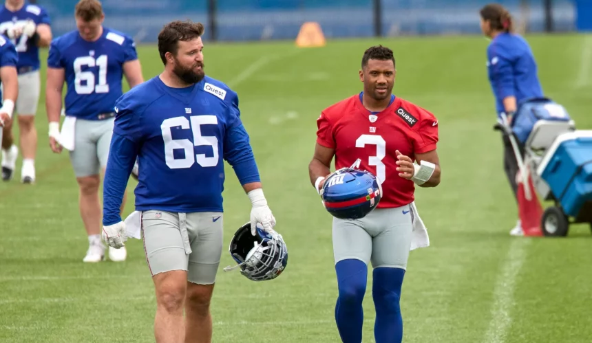 Zwei American-Football-Spieler laufen während des Giants Training auf einem Rasenplatz. Der linke Spieler trägt ein blaues Trikot mit der Nummer 65 und hält einen Helm; der rechte trägt ein rotes Trikot mit der Nummer 3 und trägt einen blauen Helm. Diese Beschreibung wurde mit der FootballR KI automatisch generiert.