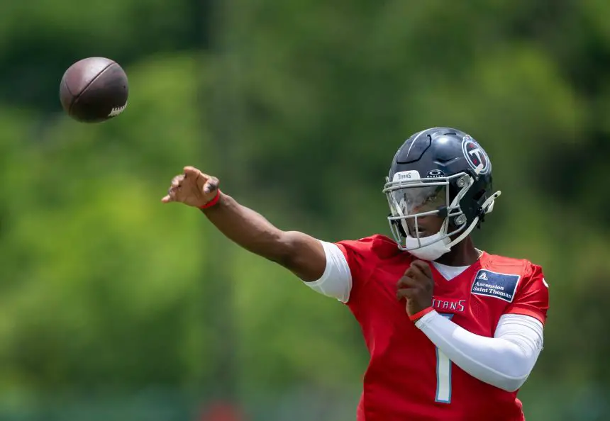 Ein Footballspieler namens Cam Ward, in einem roten Trainingstrikot und einem dunklen Helm mit der Aufschrift „TITANS“, wirft mit der rechten Hand einen Football. Er trägt einen weißen Ärmel und steht im Freien vor verschwommenen grünen Bäumen. Diese Beschreibung wurde mit der FootballR KI automatisch generiert.