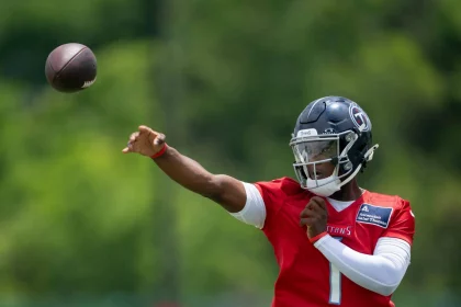 Ein Footballspieler namens Cam Ward, in einem roten Trainingstrikot und einem dunklen Helm mit der Aufschrift „TITANS“, wirft mit der rechten Hand einen Football. Er trägt einen weißen Ärmel und steht im Freien vor verschwommenen grünen Bäumen. Diese Beschreibung wurde mit der FootballR KI automatisch generiert.