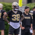 Cam Akers trägt einen Helm und die Trainingsuniform der New Orleans Saints mit der Nummer 38. Er steht mit den Händen in den Hüften. Eine weitere Person in Saints-Kleidung und mit Kappe steht neben ihm. Im Hintergrund verschwimmen die anderen Spieler und das Grün. Diese Beschreibung wurde mit der FootballR KI automatisch generiert.