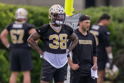 Cam Akers trägt einen Helm und die Trainingsuniform der New Orleans Saints mit der Nummer 38. Er steht mit den Händen in den Hüften. Eine weitere Person in Saints-Kleidung und mit Kappe steht neben ihm. Im Hintergrund verschwimmen die anderen Spieler und das Grün. Diese Beschreibung wurde mit der FootballR KI automatisch generiert.