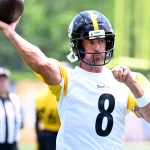 Aaron Rodgers mit schwarzem Helm und weißem Trikot mit der Nummer 8 wirft während des Spiels der Packers gegen die Steelers einen Football. Er ist mitten im Geschehen, den Blick nach vorne gerichtet, während andere Spieler und ein Schiedsrichter im Hintergrund auf einem grünen Feld verschwommen erscheinen. Diese Beschreibung wurde mit der FootballR KI automatisch generiert.