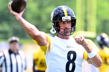 Aaron Rodgers mit schwarzem Helm und weißem Trikot mit der Nummer 8 wirft während des Spiels der Packers gegen die Steelers einen Football. Er ist mitten im Geschehen, den Blick nach vorne gerichtet, während andere Spieler und ein Schiedsrichter im Hintergrund auf einem grünen Feld verschwommen erscheinen. Diese Beschreibung wurde mit der FootballR KI automatisch generiert.