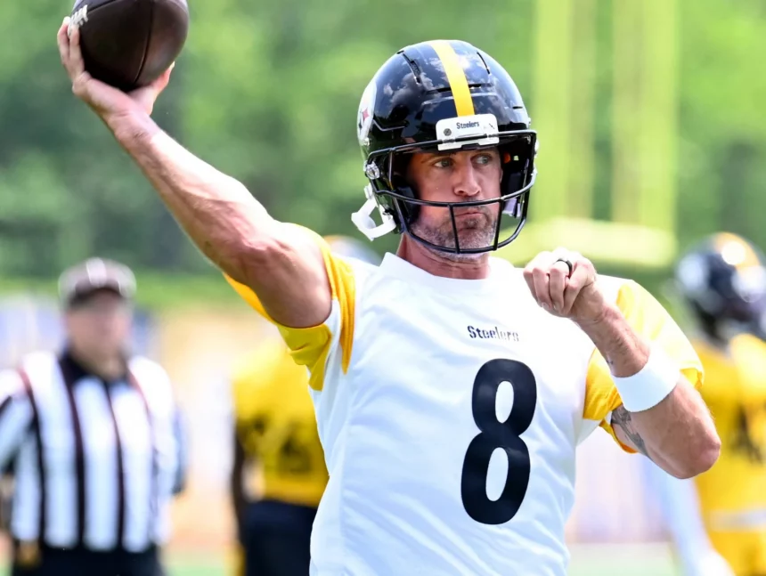 Aaron Rodgers mit schwarzem Helm und weißem Trikot mit der Nummer 8 wirft während des Spiels der Packers gegen die Steelers einen Football. Er ist mitten im Geschehen, den Blick nach vorne gerichtet, während andere Spieler und ein Schiedsrichter im Hintergrund auf einem grünen Feld verschwommen erscheinen. Diese Beschreibung wurde mit der FootballR KI automatisch generiert.